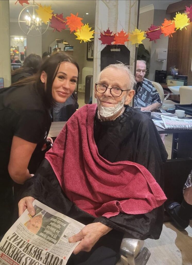 male resident sat in hairdressing chair with a newspaper in his hand having his beard shaved and smiling with the hairdresser