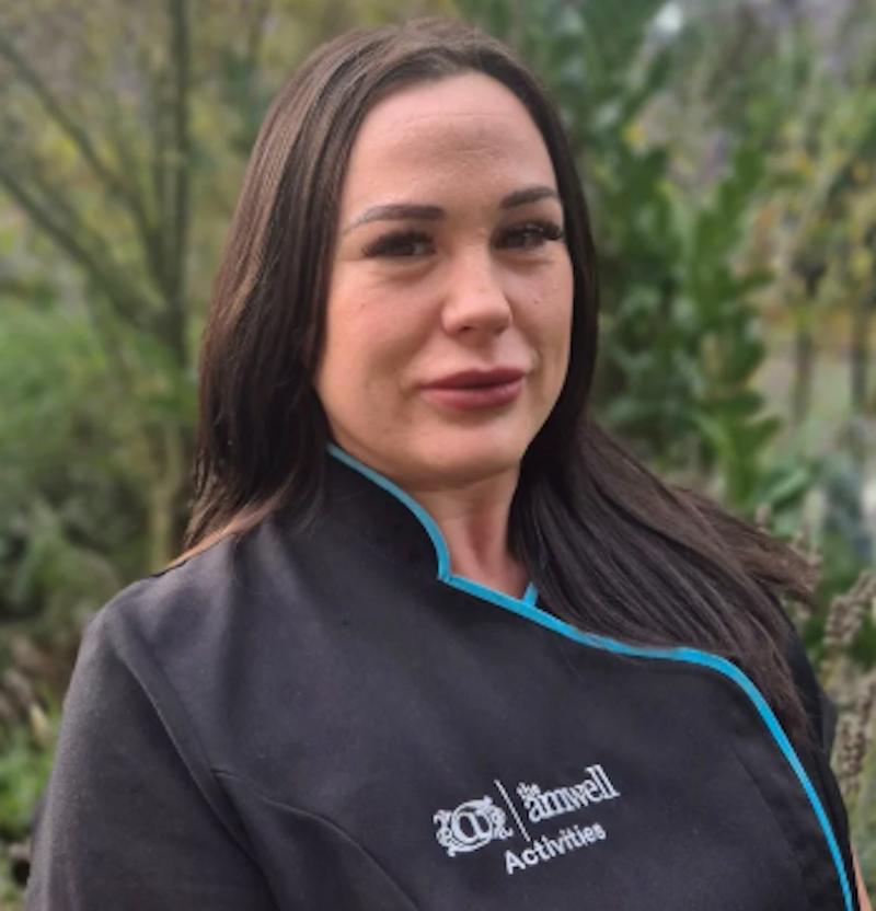 Woman with brown hair in a black and blue activities coordinator uniform having a headshot taken outside for the team page