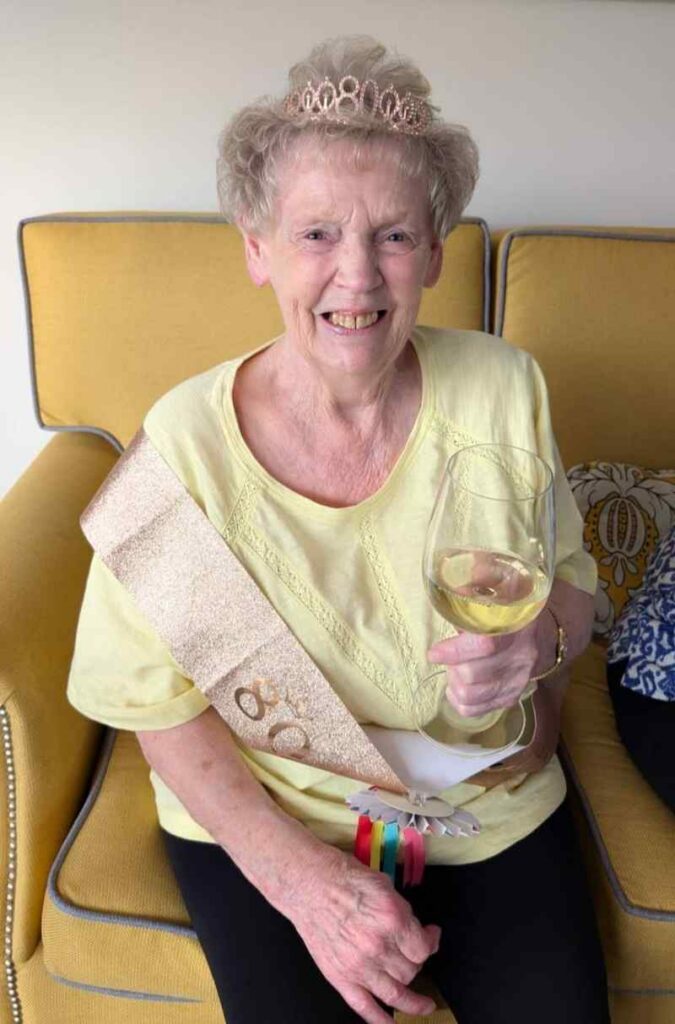 one of our residents wearing a yellow top, a sash and a crown sat down smiling with a glass of wine in her hand