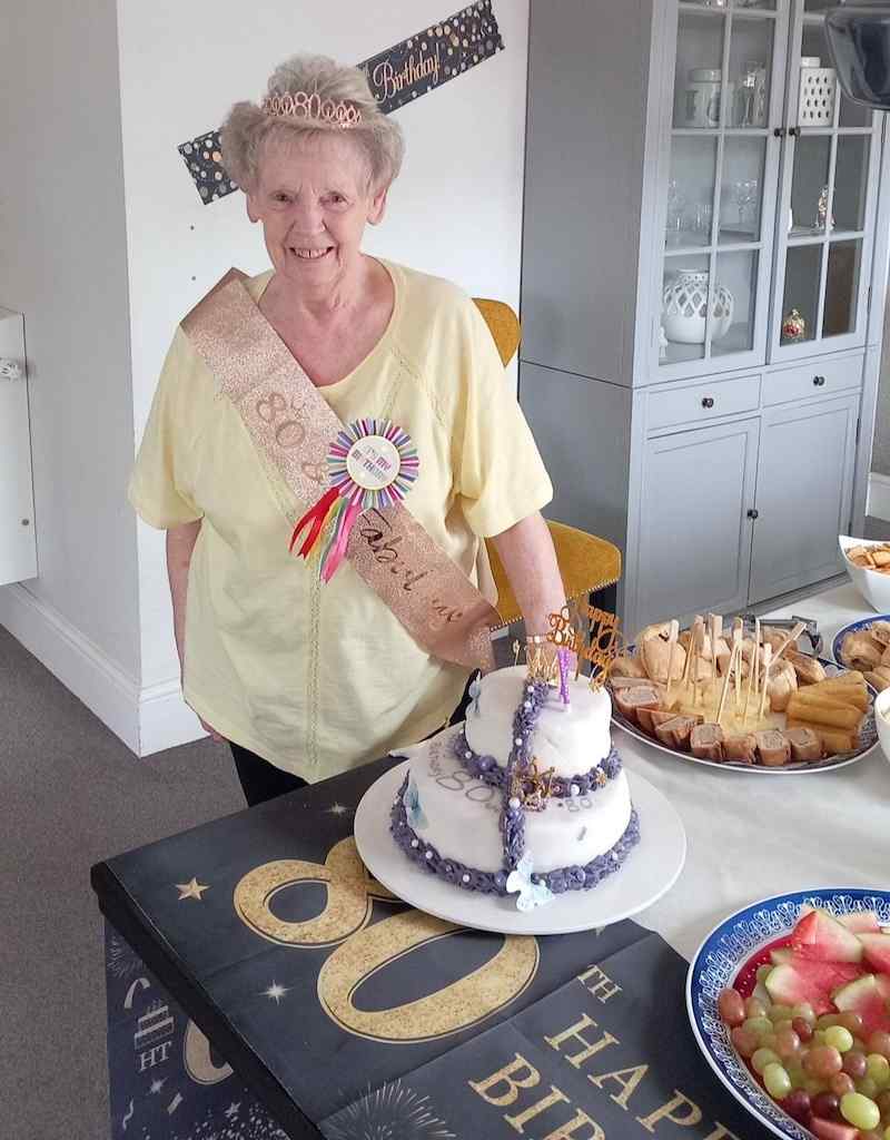 one of our residents wearing a yellow top, a sash and a crown smiling in front of a birthday cake