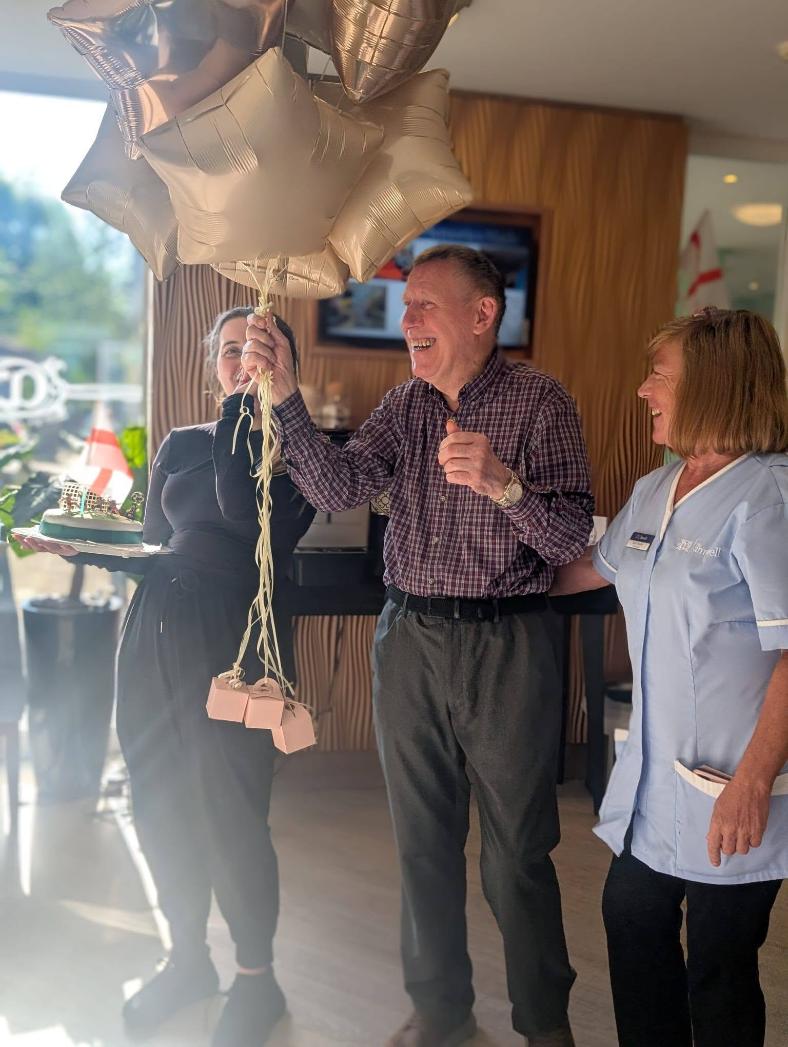 an older man with care staff members holding birthday balloons and cake